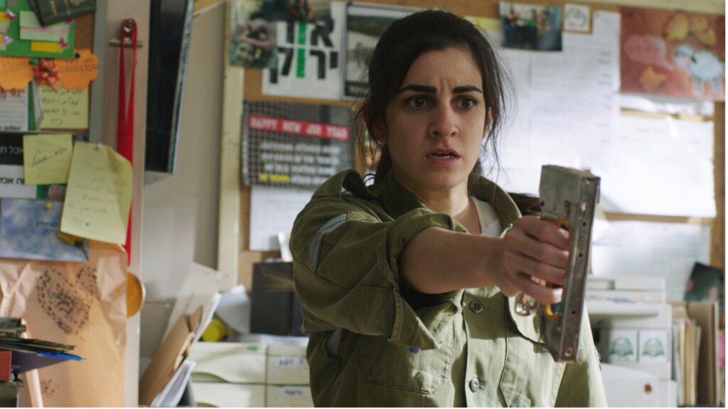 A woman in a green military shirt holds a staple gun in an outstretched hand. Behind her are noticeboards and files.