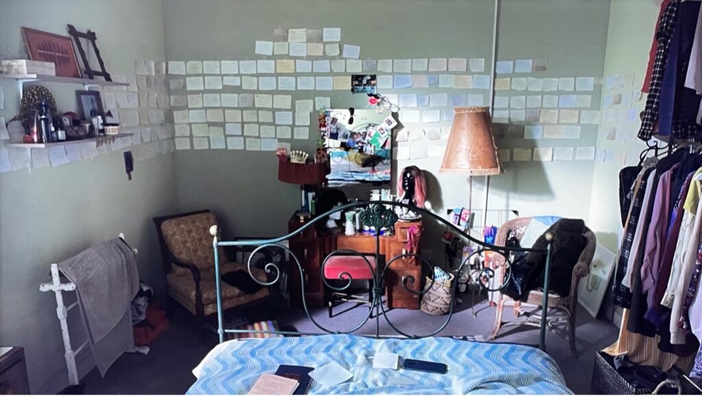 A bedroom viewed from the bed. Sticky notes cover the walls; there are also rails of clothes, two chairs, a dressing table, and shelves.