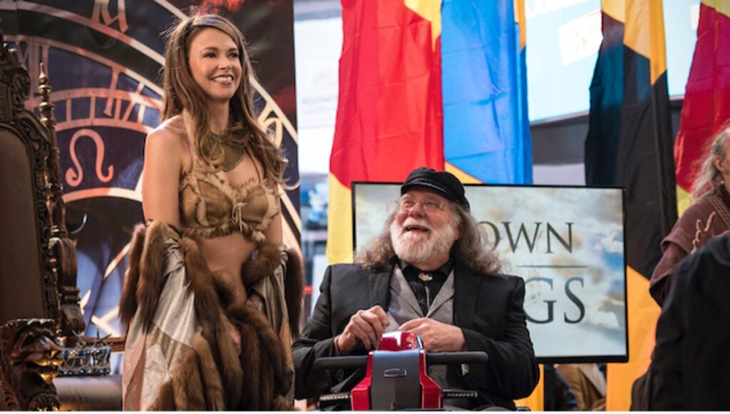 Liza stands to the left of Empirical author, L. L. Moore, who sits on a motorised scooter. She is dressed as his character Princess Pam Pam in a parodically sexy bikini made of fur and animal skins. He is looking at her adoringly.