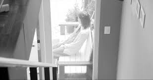 Still from Hong Sang-soo's film Grass, shows a person sitting on a porch, shot from inside and up a flight of stairs.